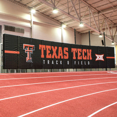 A large black track and field sideline banner with 'TEXAS TECH' in Red, the Texas Tech 'T' logo, and 'TRACK & FIELD' in white, installed on an indoor running track.